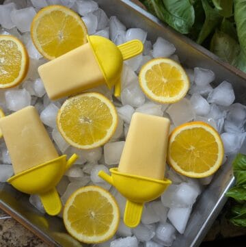 picture of orange Julius popsicles surrounded by orange slices, resting on a bed of ice in a stainless steel pan, on a wooden cutting board and counter with green plants