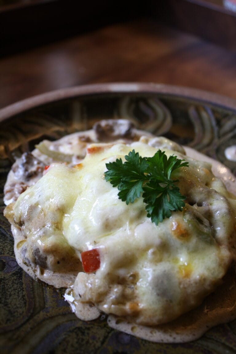 A plated casserole dish, sitting on a dark brown plate and cutting board, covered in melted cheese and a sprig of parsley.