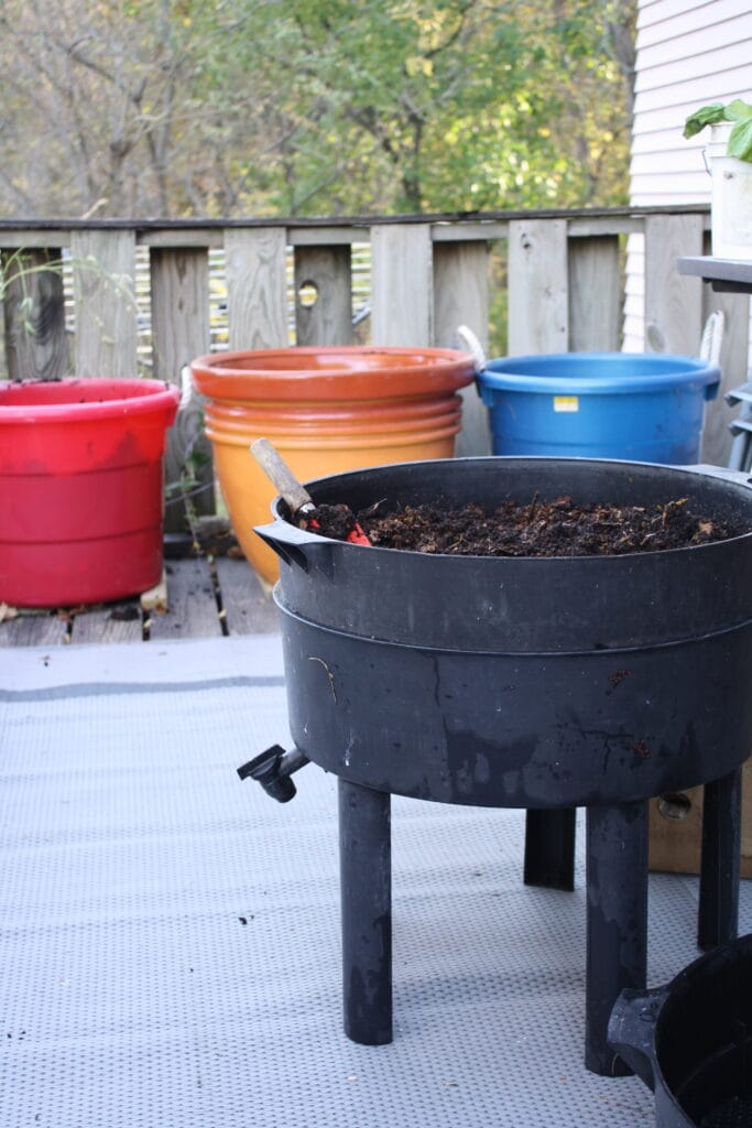 a worm bin on a deck with pots behind it in red, orange and blue, waiting to be amended with worm castings so the bin can be brought inside for winter