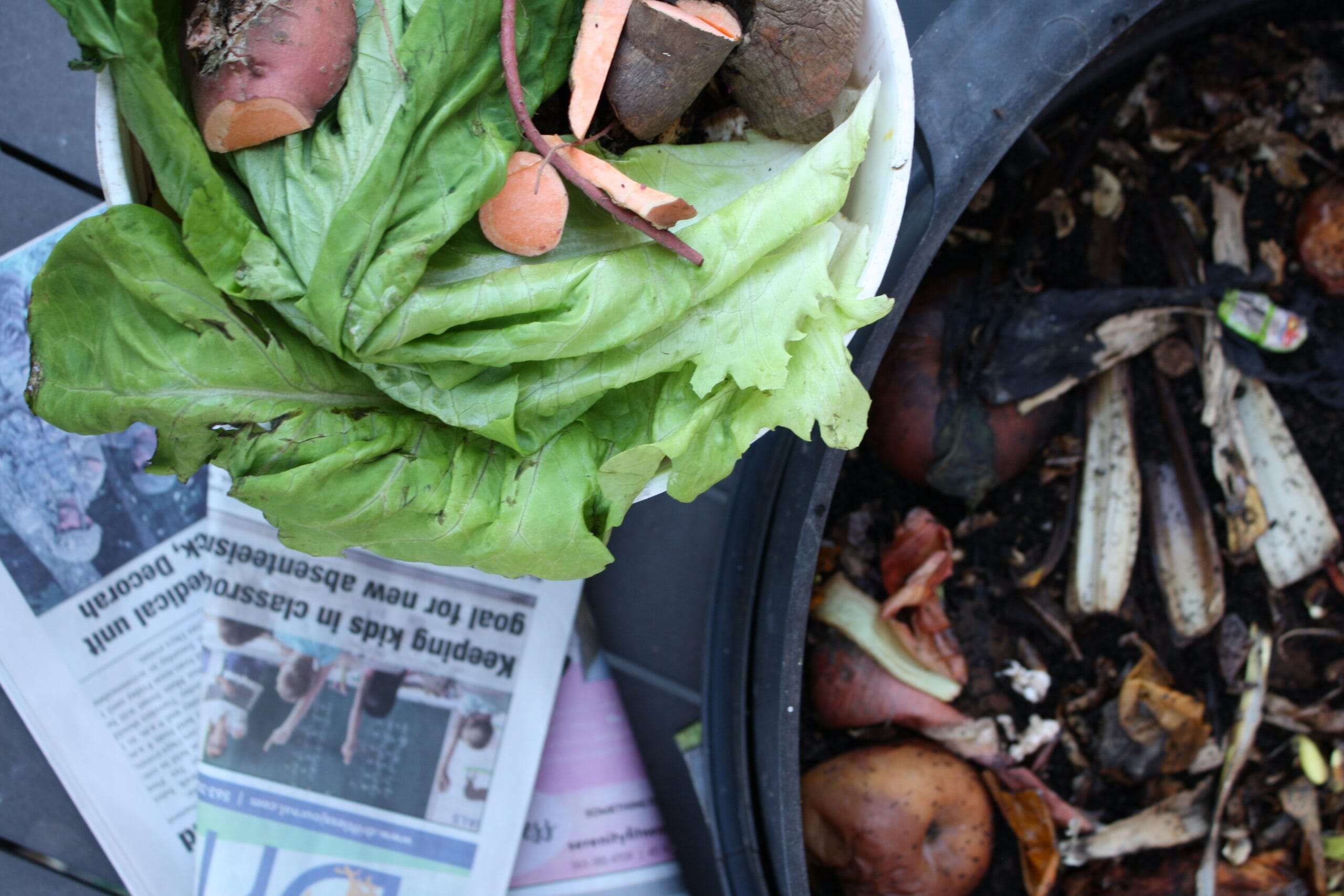 a small bucket of fresh food scraps - wilted lettuce, sweet potato ends, etc - next to an almost composted worm bin later and a pile of newspaper, getting ready to feed to the worms.