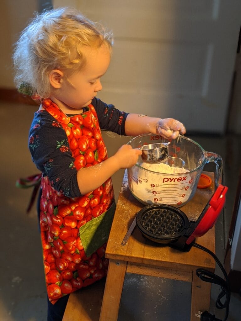 A little girl in an apron, measuring ingredients into a big glass bowl next to a mini waffle maker, learning Simple Kitchen Habits for Beginning Homemakers