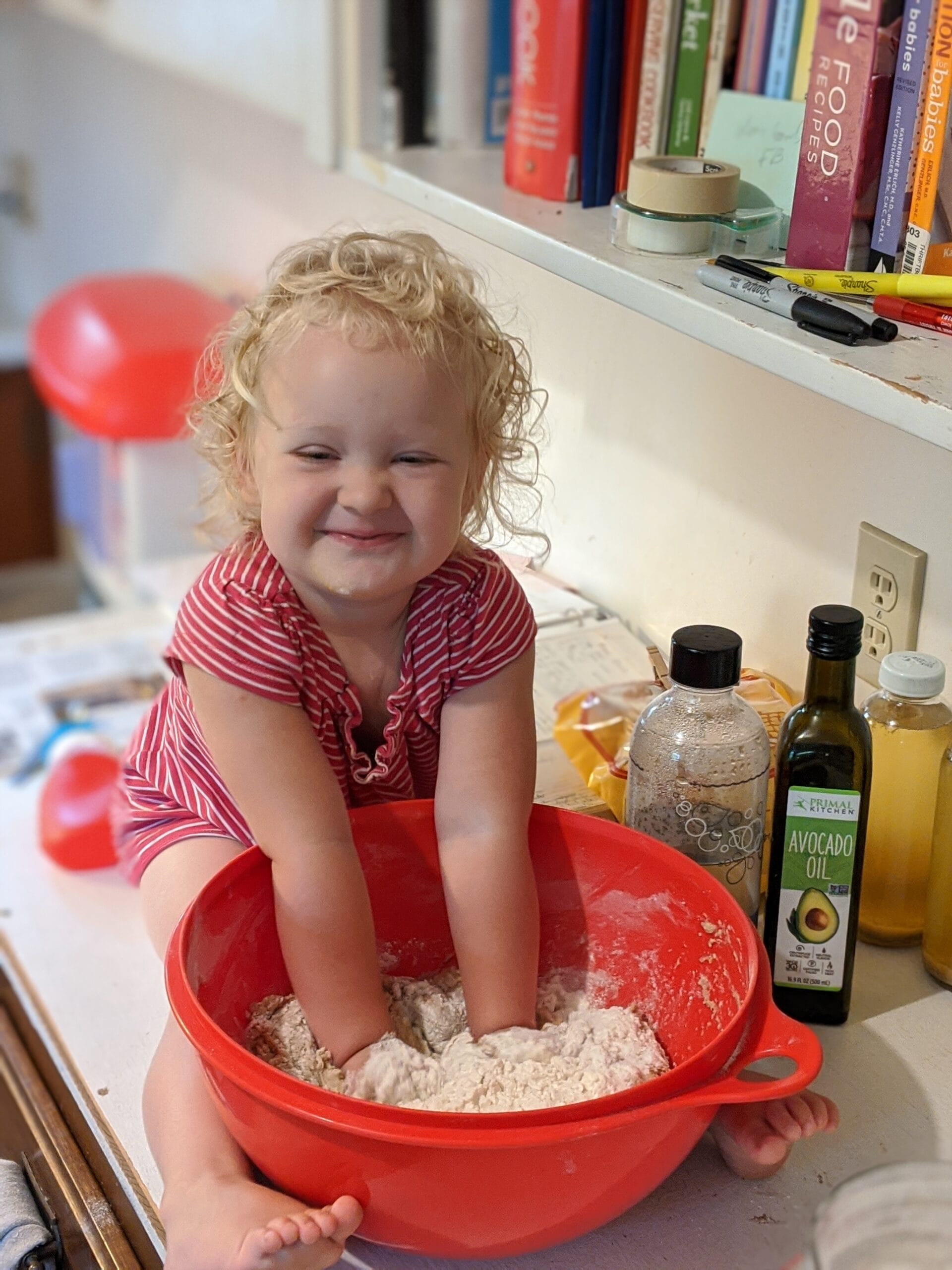 a little girl sitting on the kitchen counter, pushing her hands into a big bowl of dough, with a smile on her face