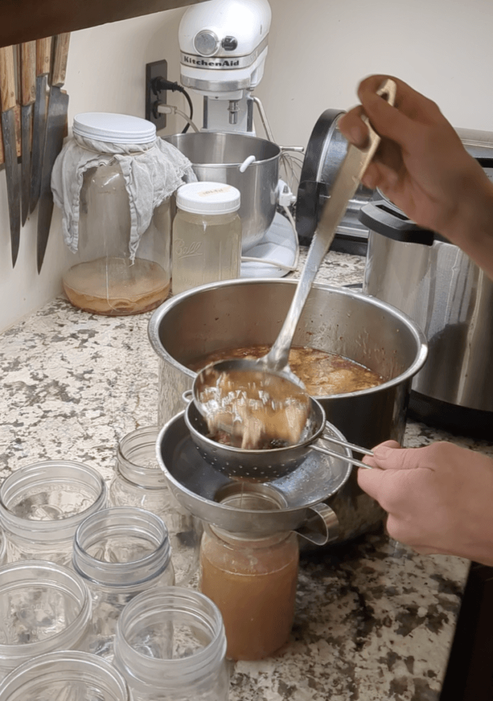 A caucasian woman is ladling bone broth into jars through a strainer on her kitchen counter.