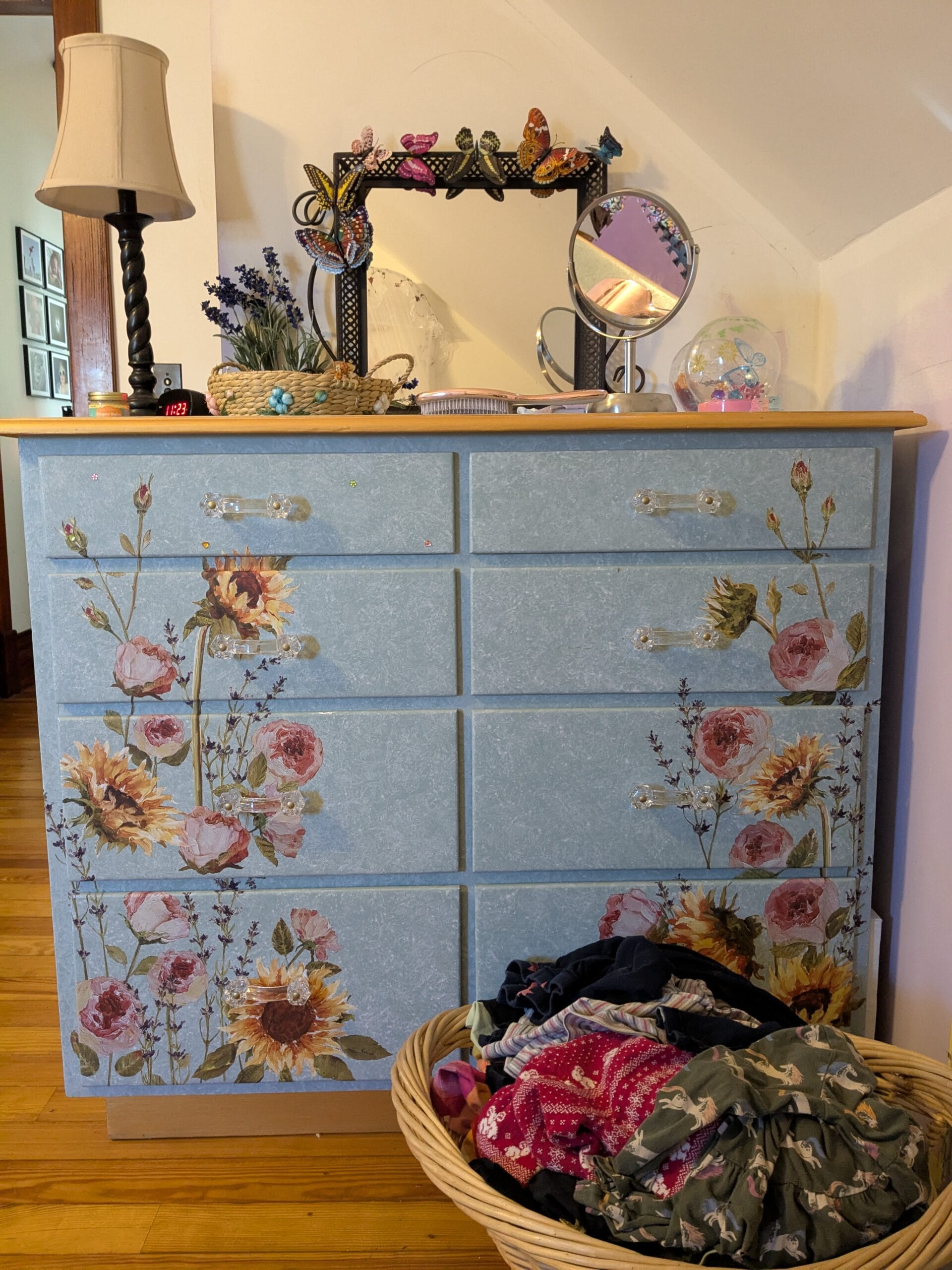 When I stopped folding my kids' laundry, I set a wicker laundry basket full of clothes on the floor in front of a dresser decorated with beautiful, colorful flowers with a lamp and mirror on top.