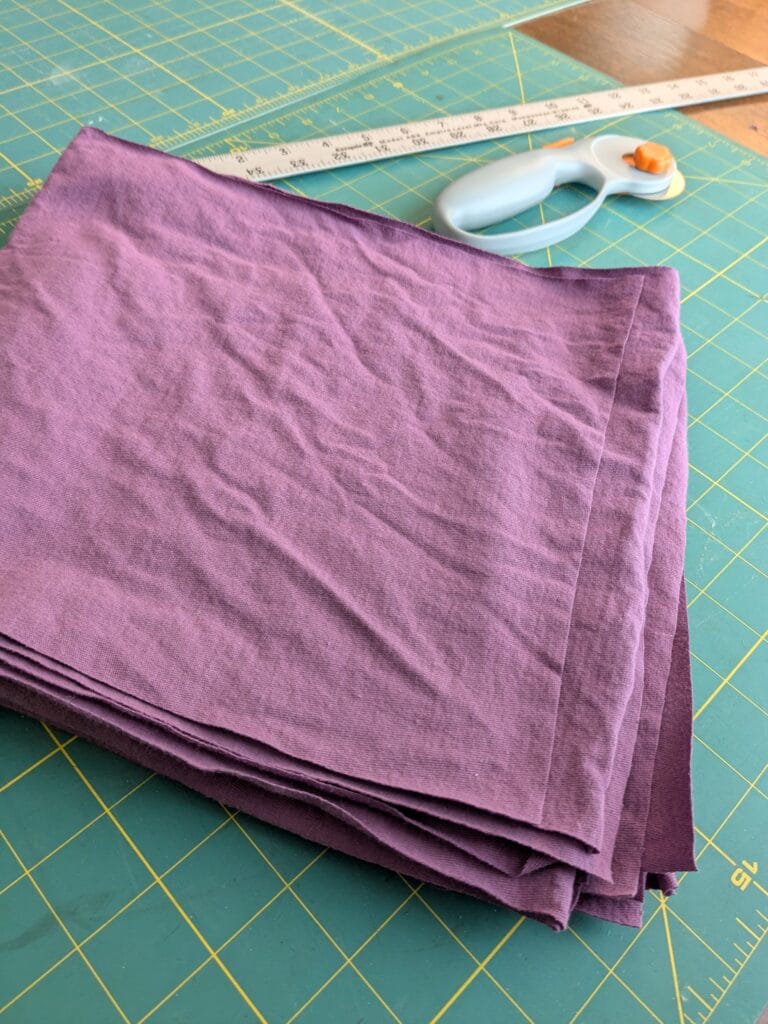 a pile of squares cut out of a thin knit berry colored fabric with measuring and cutting tools in the background