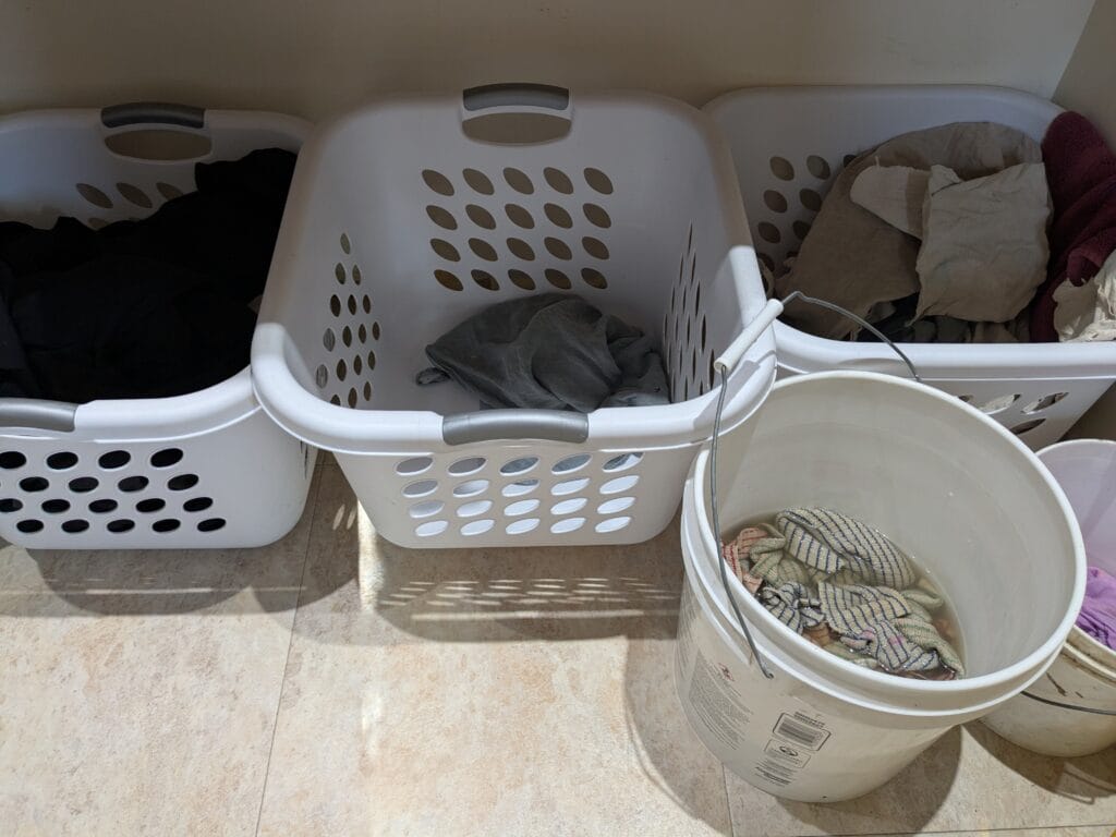 Clothes and rags sorted into three square laundry baskets and two soaking buckets sitting on the floor.