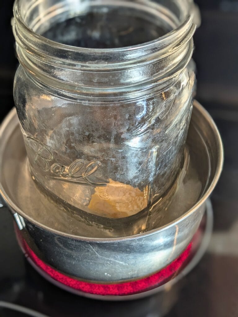 a small pot sits on a red hot burner with a quart mason jar sitting inside - like a double boiler. Inside the glass jar is a small chunk of beeswax, melting.