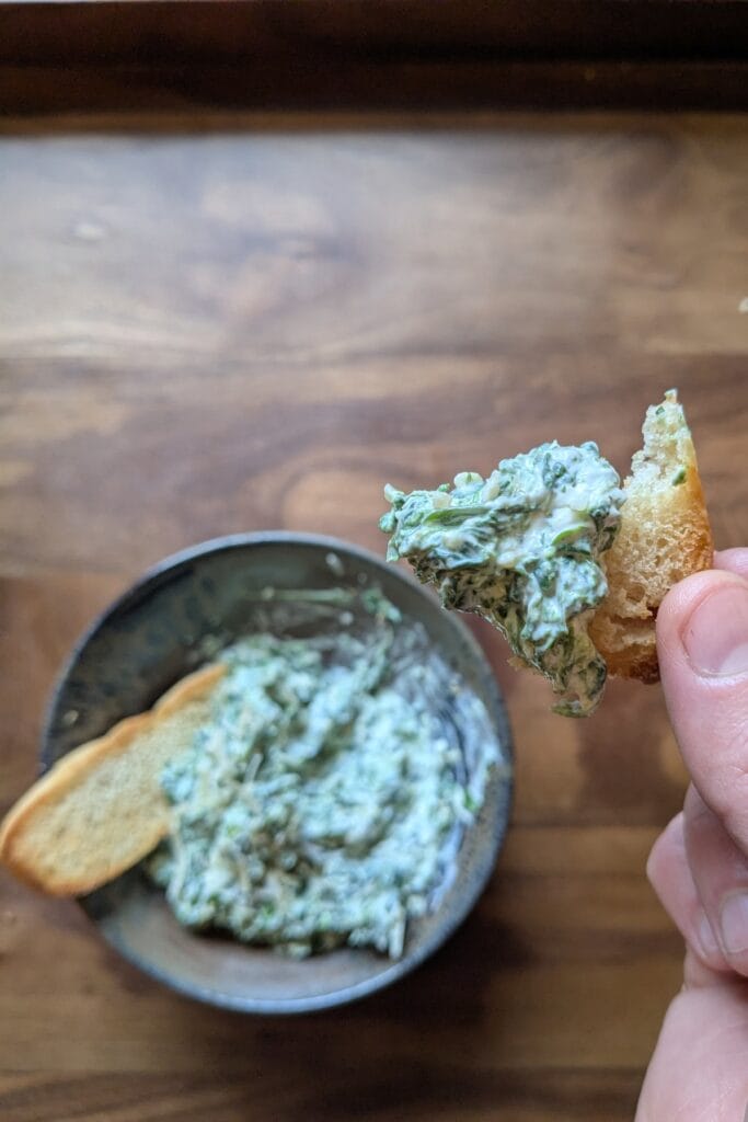 A dark ceramic bowl holding a big scoop of homemade spinach dip. There is one thin toasted slice of sourdough bread sticking up out of the dip and a hand holding another one, bitten, with a new scoop full of the dip.