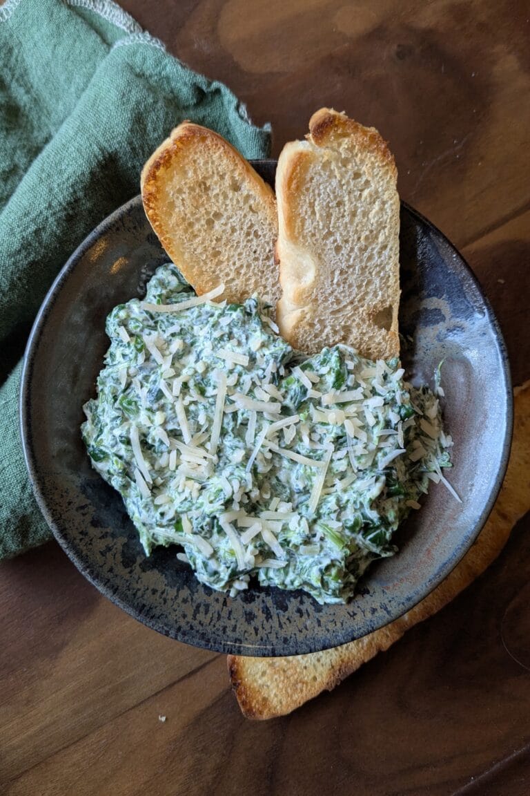 A dark ceramic bowl holding a big scoop of homemade spinach dip. There are two thin toasted slices of sourdough bread sticking up out of the dip.