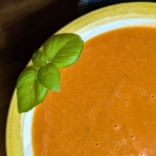 Yellow stripe bowl of tomato soup with fresh basil garnish, resting on a wooden cutting board with a navy blue linen napkin beside it.