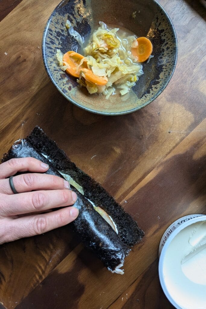 a caucasian hand is almost done rolling up a piece of seaweed covered in cream cheese, sliced meat, and sauerkraut laying on a dark wooden tray, surrounded by other ingredients to roll up in it.