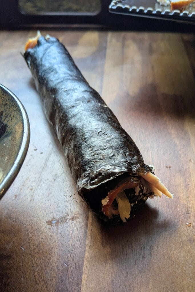 a rolled up piece of seaweed stuffed with cream cheese, sliced meat, and sauerkraut laying on a dark wooden tray, surrounded by other ingredients to roll up in it.