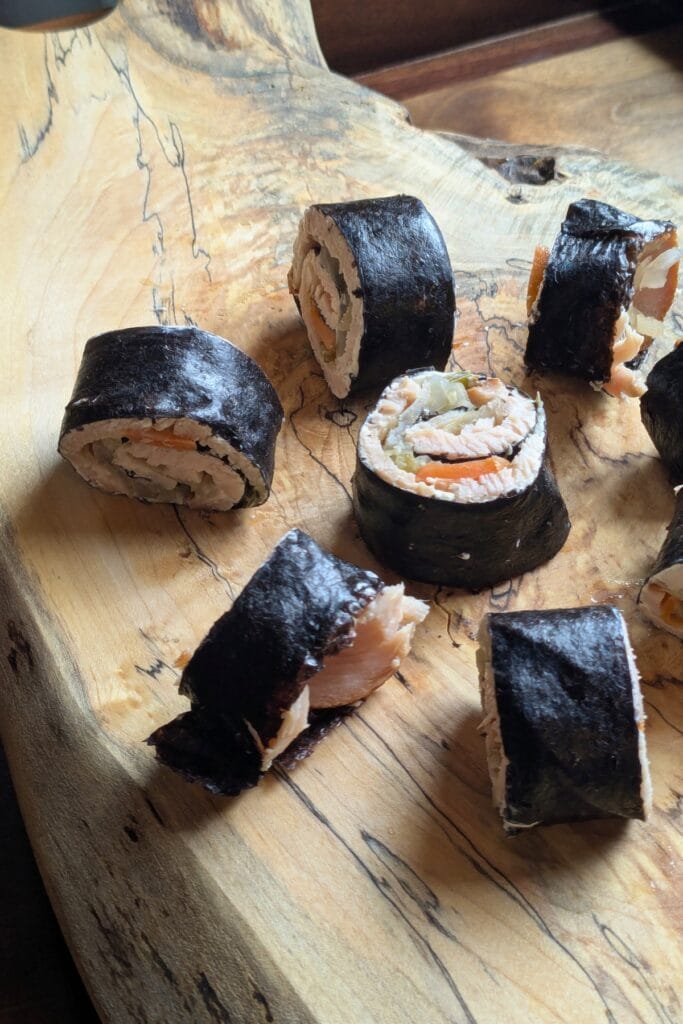 a beautiful wooden cutting board holding one whole half and one cut up half of a seaweed roll-up.