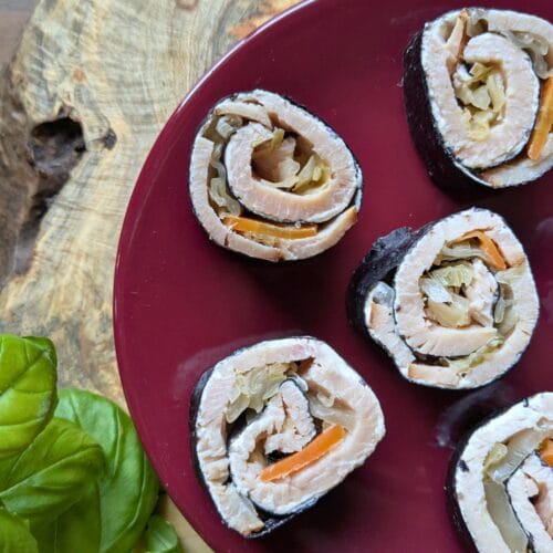 a cutting board with a small burgundy plate with sliced up ham and cheese seaweed roll-ups on it with basil leaves around it