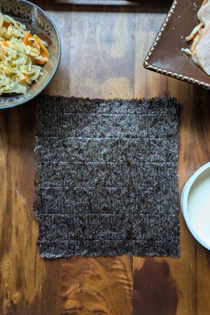 a piece of seaweed laying on a dark wooden tray, surrounded by other ingredients to roll up in it.