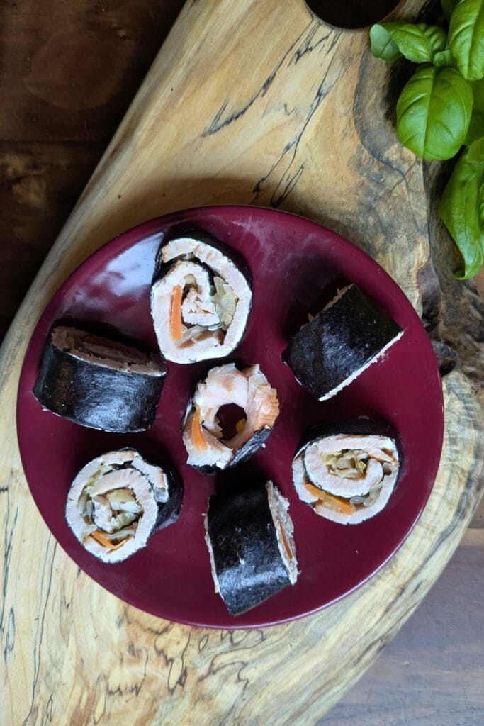 a cutting board with a small burgundy plate with sliced up ham and cheese seaweed roll-ups on it with basil leaves around it