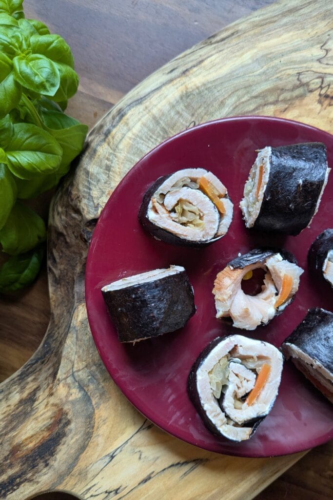 a cutting board with a small burgundy plate with sliced up meat and cheese seaweed roll-ups on it with basil leaves around it