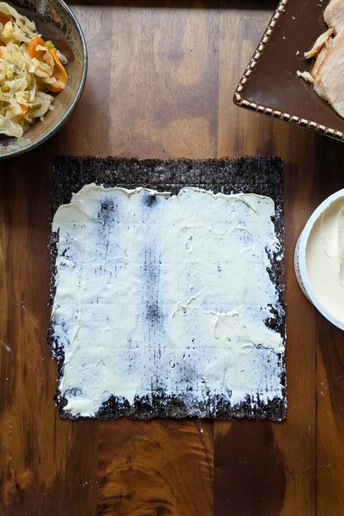 a piece of seaweed covered in cream cheese laying on a dark wooden tray, surrounded by other ingredients to roll up in it.