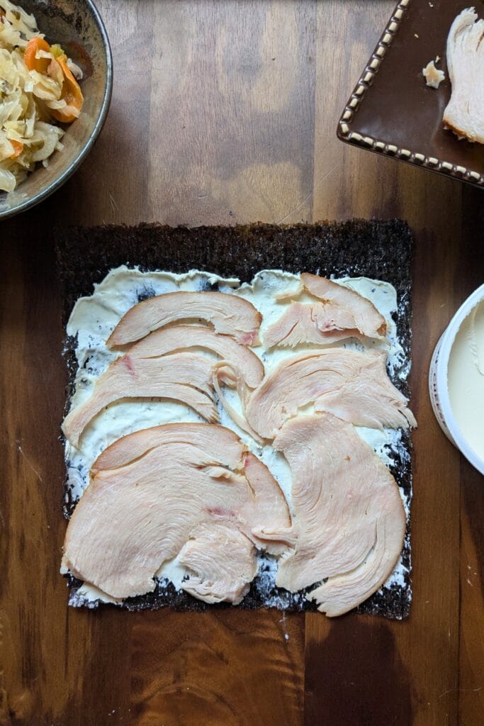 a piece of seaweed covered in cream cheese and sliced meat laying on a dark wooden tray, surrounded by other ingredients to roll up in it.