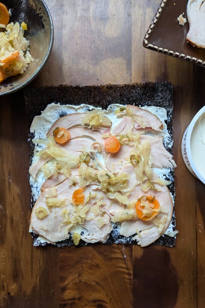 a piece of seaweed covered in cream cheese, sliced meat, and sauerkraut laying on a dark wooden tray, surrounded by other ingredients to roll up in it.