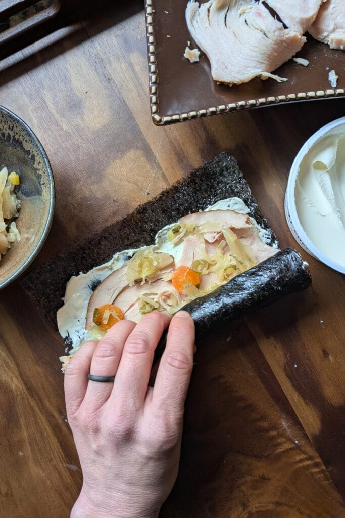 a caucasian hand is rolling up a piece of seaweed covered in cream cheese, sliced meat, and sauerkraut laying on a dark wooden tray, surrounded by other ingredients to roll up in it.
