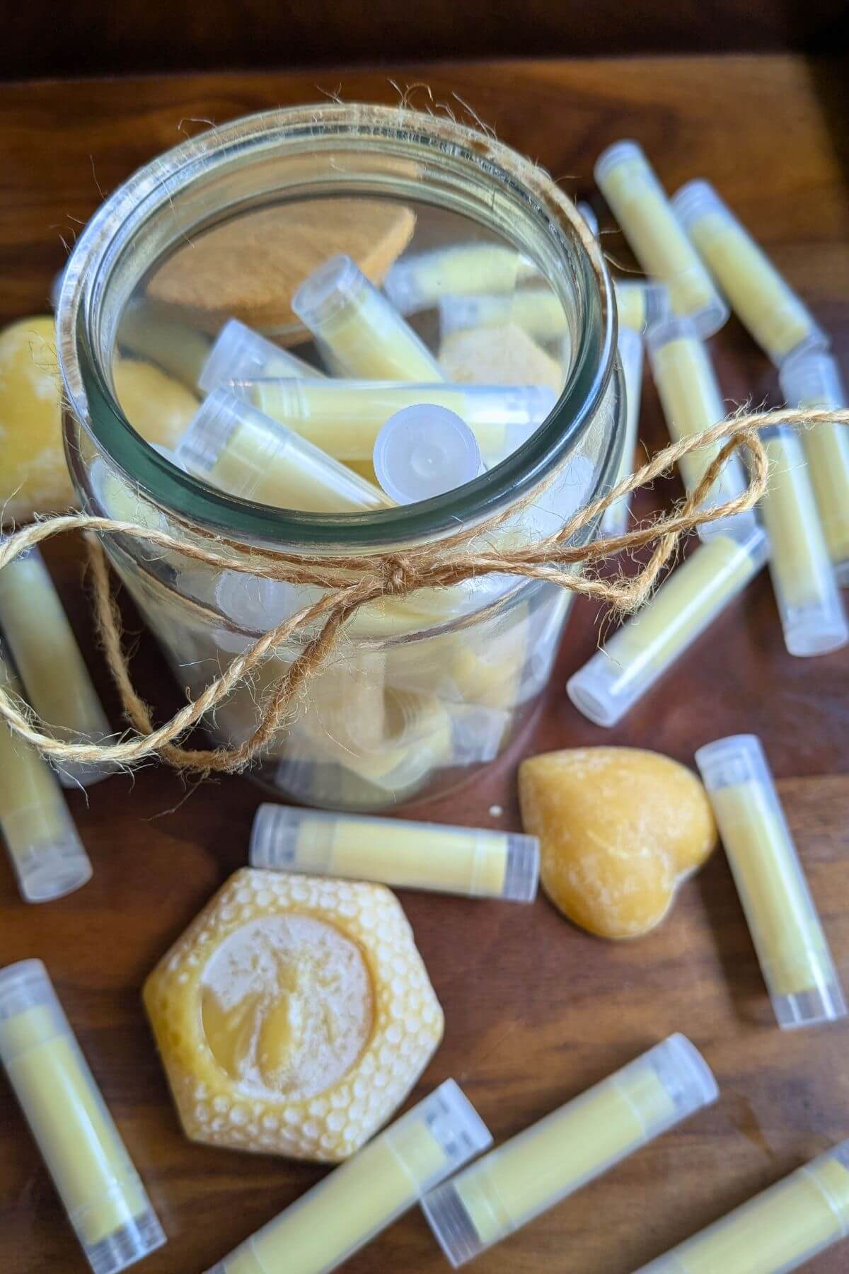 A glass jar filled with clear tubes of homemade lip balm. A string of twine is tied around the tip and a wooden lip rests against the side. more tubes of homemade chapstick and cakes of beeswax are scattered round the jar and sitting on a wooden tray.