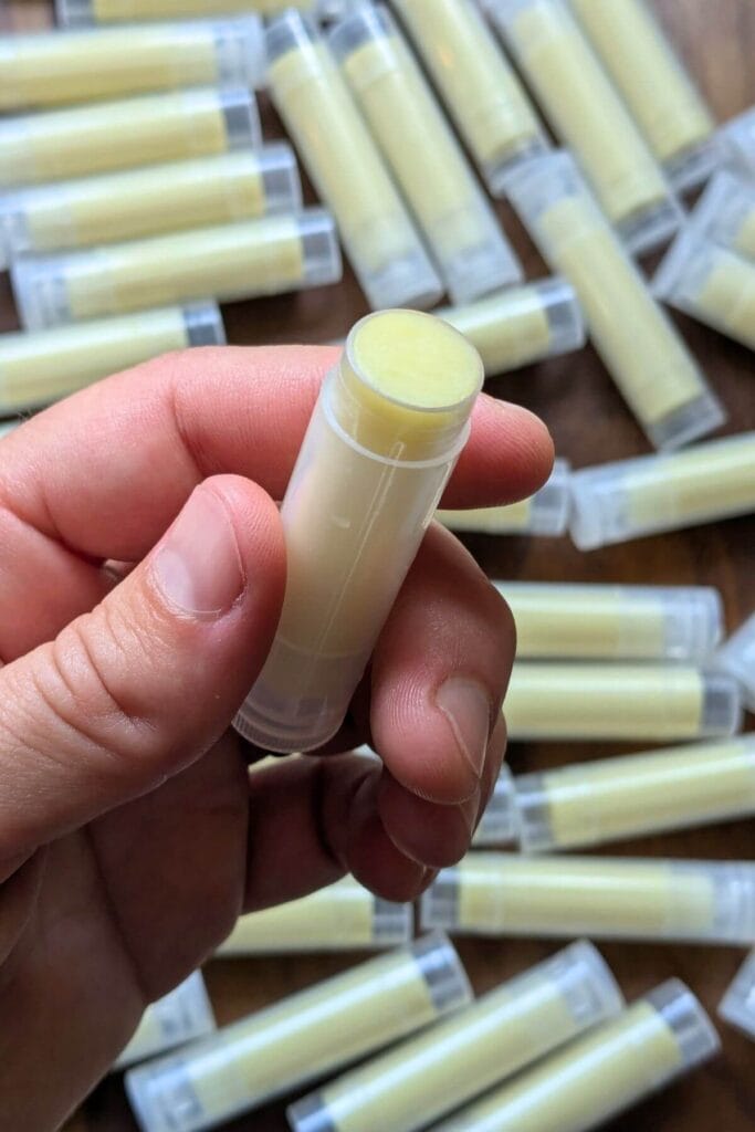 tubes of homemade chapstick are scattered round the jar and sitting on a wooden tray. A hand is holding one opened tube up to the camera.