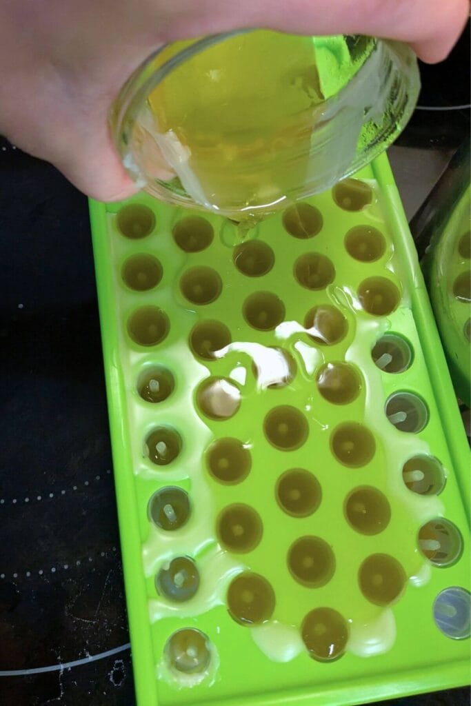 A jar of melted homemade natural lip balm mixture is held by a hand pouring it into the empty tubes that fill a bright green filling tray.