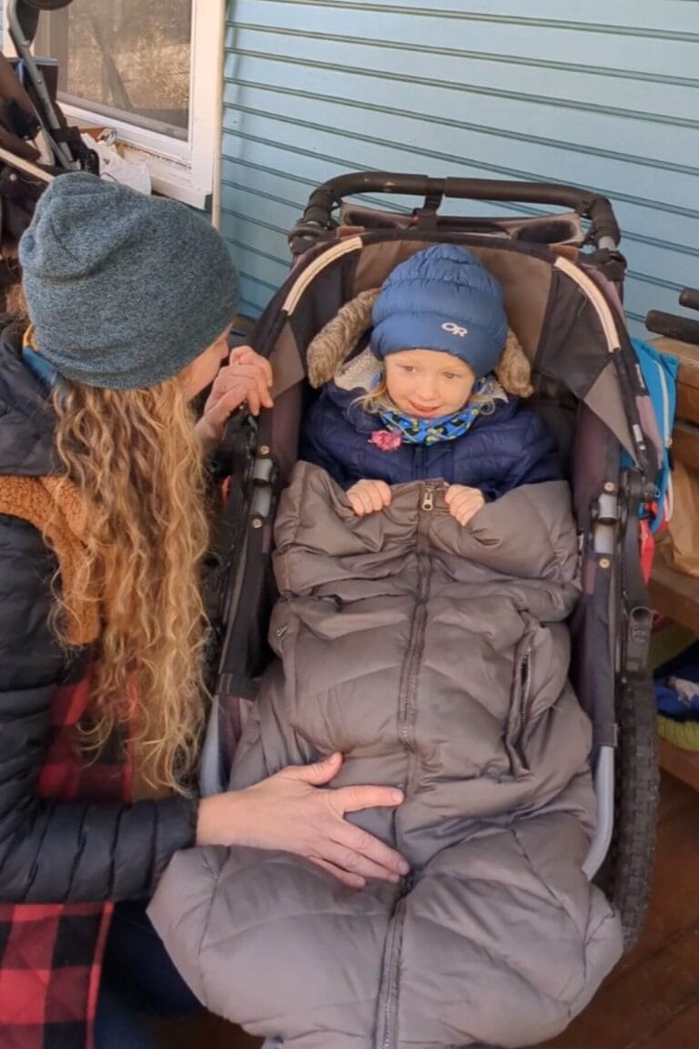 A mom crouching down by her happy little one in a Bob Jogging Stroller with an up-side-down adult coat zipped up around the toddler to keep her warm on a winter stroller walk.