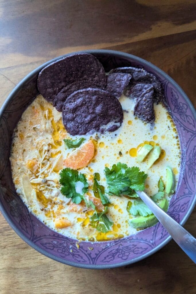 A purple bowl surrounded by purple tortilla chips, filled with white chicken soup with no beans, garnished with cilantro and avocado