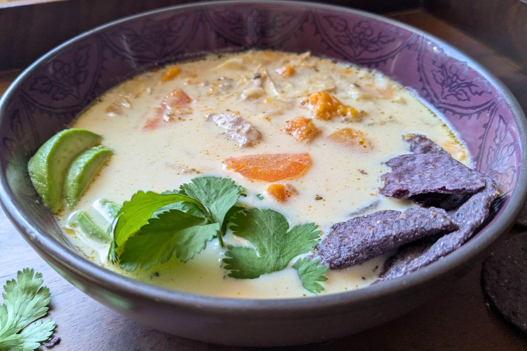A purple bowl surrounded by purple tortilla chips, filled with white chicken soup with no beans, garnished with cilantro and avocado