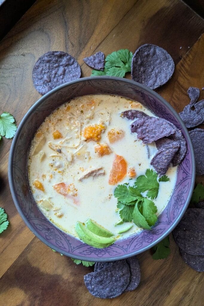 A purple bowl surrounded by purple tortilla chips, filled with white chicken soup with no beans, garnished with cilantro and avocado