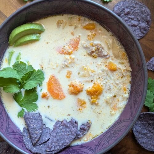 A purple bowl surrounded by purple tortilla chips, filled with white chicken soup with no beans, garnished with cilantro and avocado