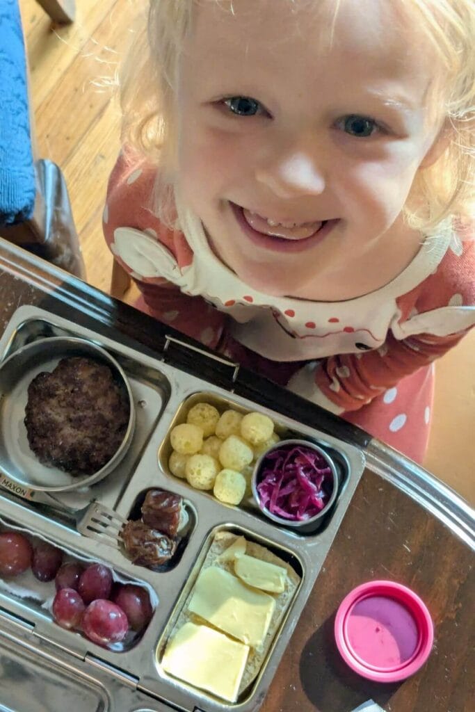 A cute little kid smiling up at the camera while she sits in front of her balanced school lunch packed in stainless steel PlanetBox Rover lunch box