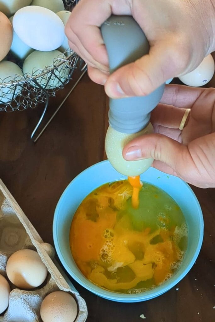 Hands using a turkey baster bulb to blow out the inside of a raw egg into a bowl using a no-mouth method for decorating eggs