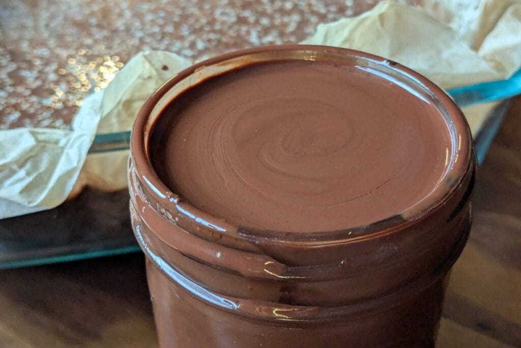 melted chocolate chips and coconut oil in a jar for a homemade dark chocolate magic shell recipe. A pan of bars are in the background with the magic shell covering them.