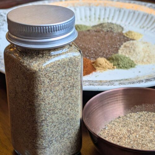 Ingredients for homemade chophouse seasoning including smoked salt, garlic, herbs and spices and Homemade chophouse seasoning in a glass jar beside them.